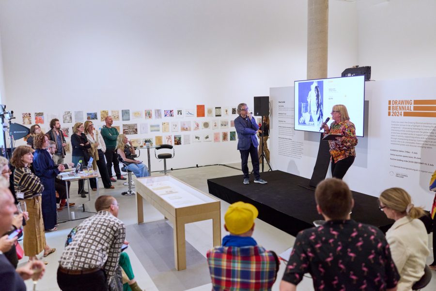 A group of people in a gallery exhibition watch a woman onstage speaking in a microphone